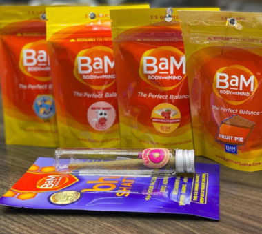 Assorted colorful packaged cannabis products and a small glass vial with cannabis inside displayed on a surface