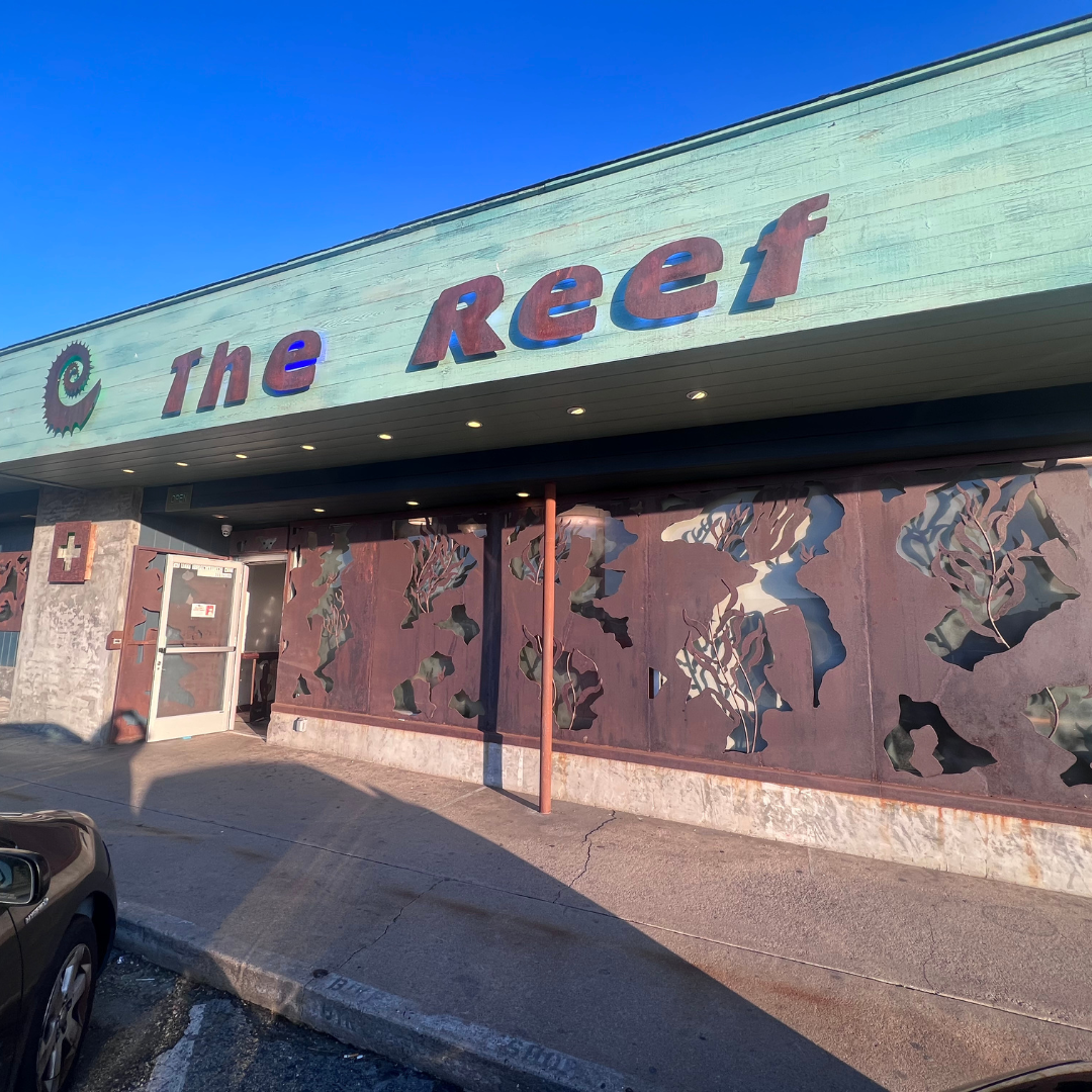 Street-level view of a building with a sign that reads 'The Reef' and decorative ocean-themed wall panels beneath a teal awning.