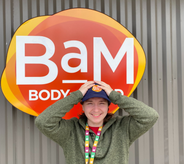 A smiling person wearing a green sweater and a blue cap standing in front of a colorful BaM Body sign