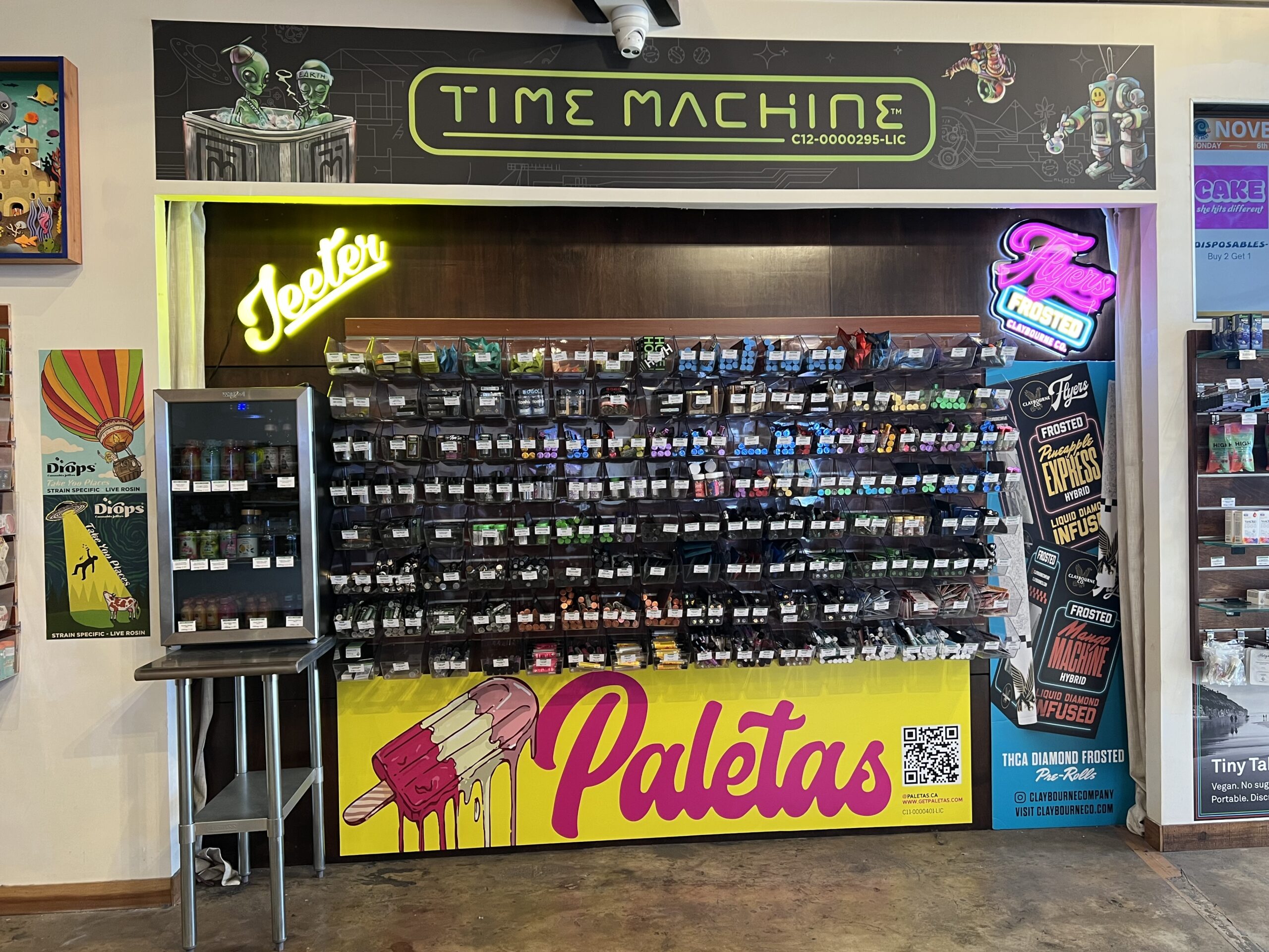 A colorful retail display with various packaged products, neon signs, and a large 'Paletas' ice cream advertisement