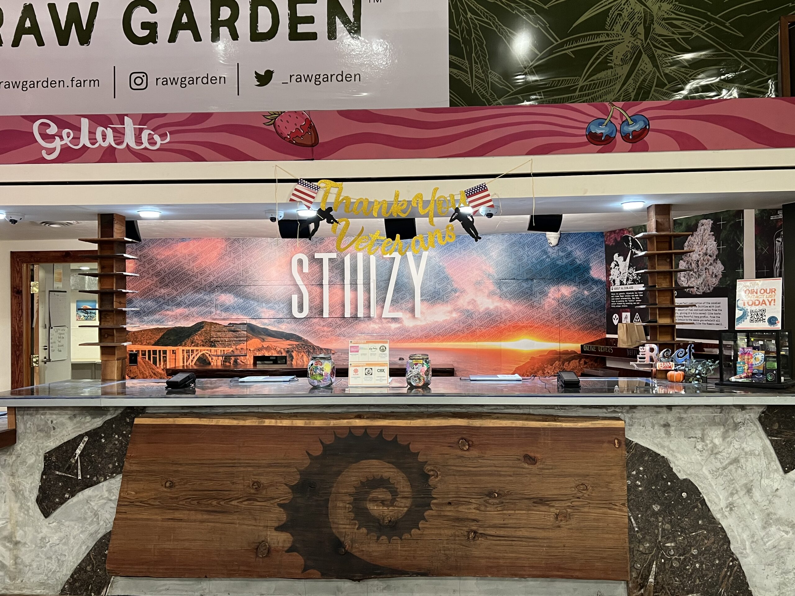 Counter area with a sunset mural, wooden front panel with a spiral sun design, and 'Thank You Veterans' and 'STIIIZY' signage above