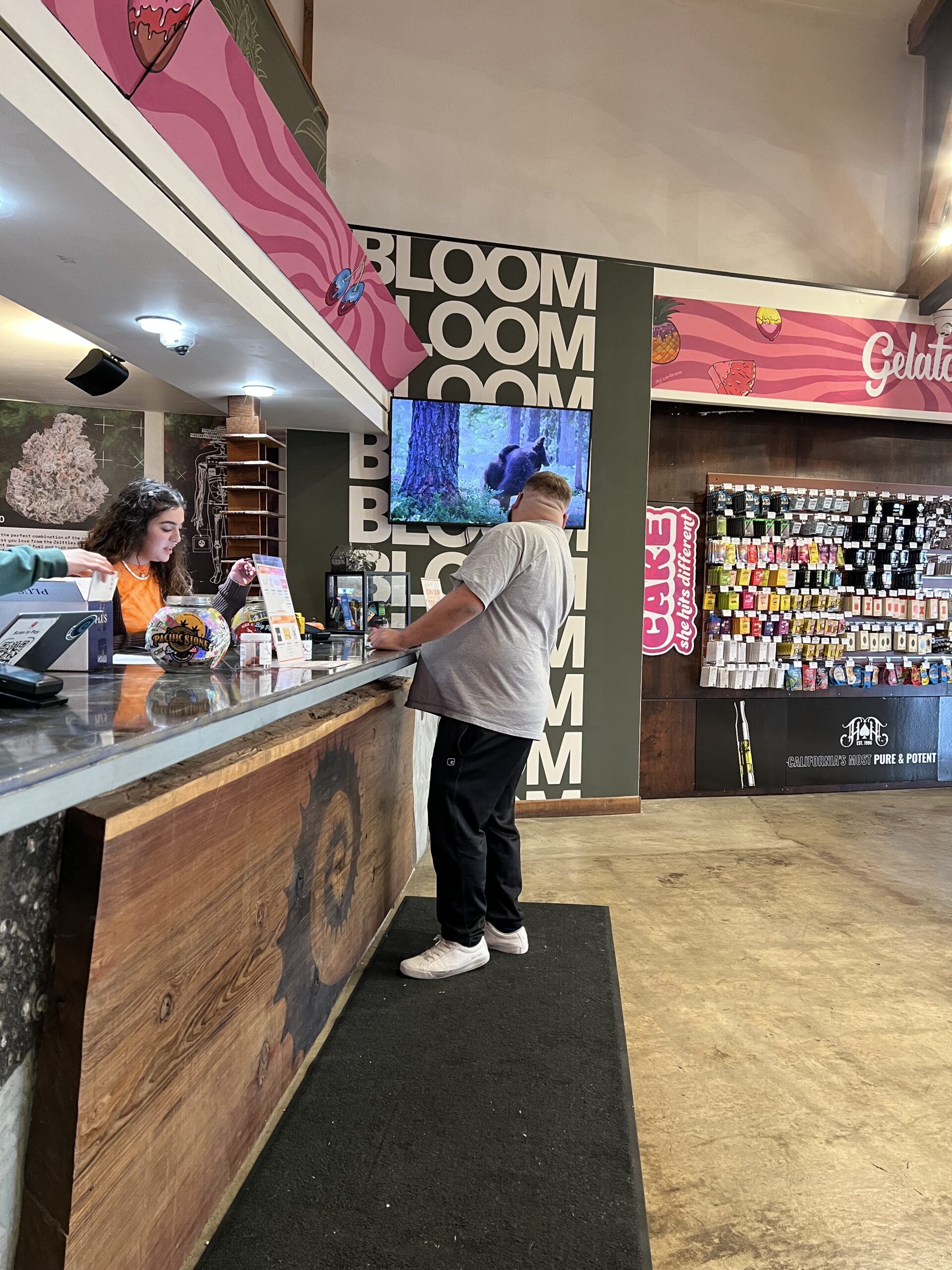 A man wearing a cap stands at a counter while a woman behind the counter assists him in a store with colorful signage and product displays